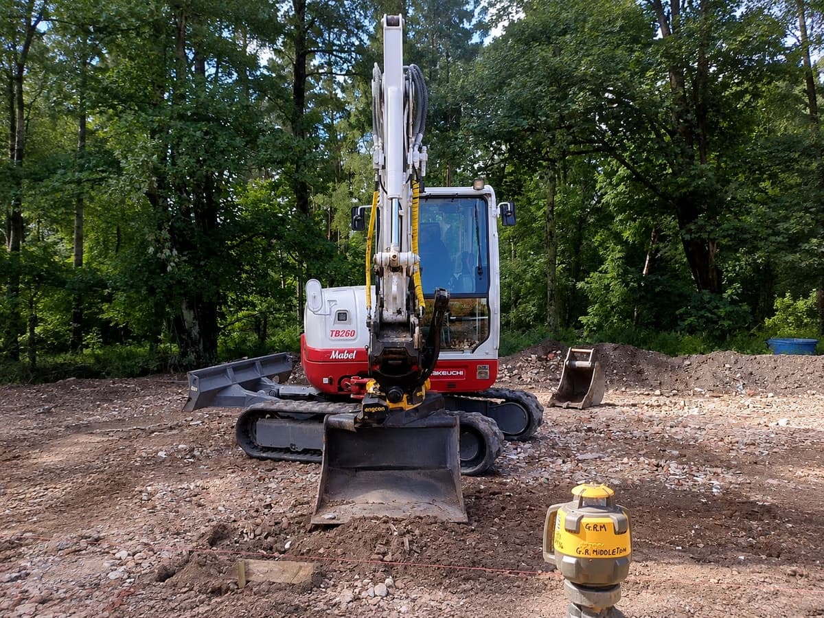 Gabriel's Takeuchi digger on a cleared plot, footings set out with string lines and a laser level in the foreground