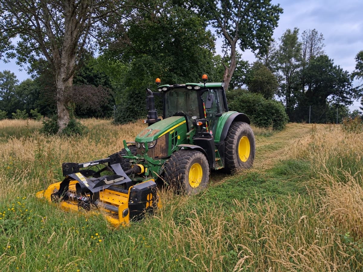 A tractor with a flail mower topping long grass under the trees, the cut ground behind it